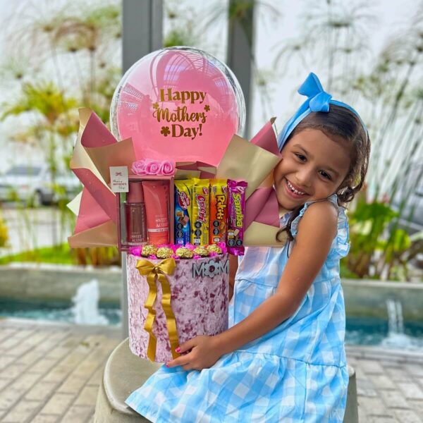 Niña abrazando caja rosa de regalo para mamá con chocolates y globo rosado.