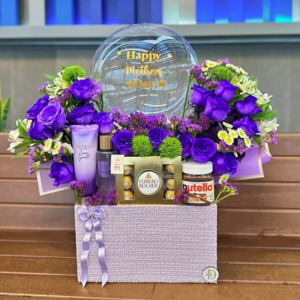 Arreglo floral lila con chocolates Ferrero, Nutella y cremas Victoria’s Secret. Caja lila con rosas violetas, astromelias blancas, margaritas, Ferrero Rocher, Nutella y set de cremas Victoria's Secret Love Spell, decorada con globo “Happy Mother’s Day”.